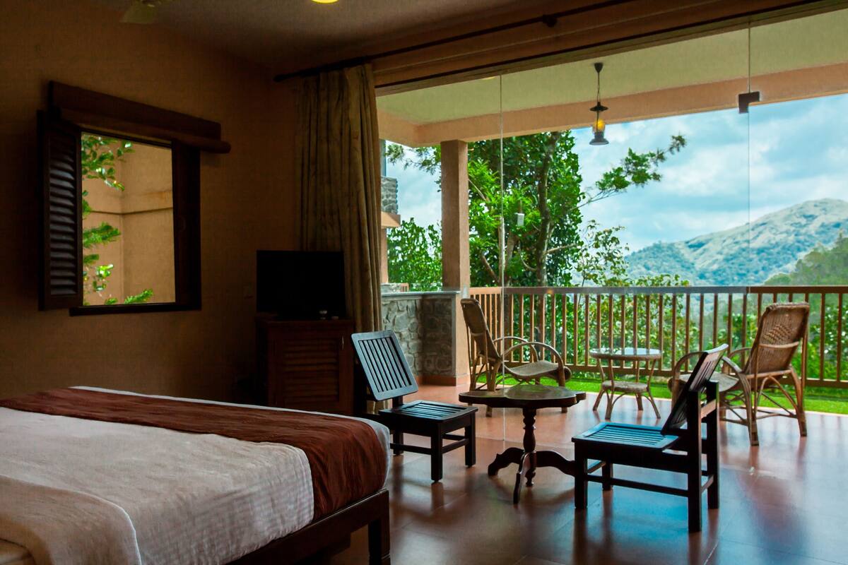 A bedroom with a bed, a TV, a balcony with chairs and a table, and a view of greenery and mountains.