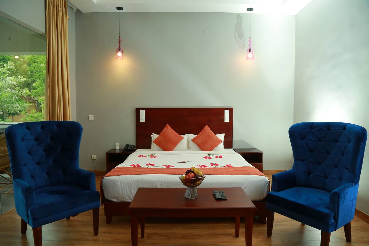 A hotel room with a large bed, two blue armchairs, a wooden nightstand, a small table with a fruit bowl, and a view of greenery through the window.