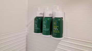 Tre flaconi di Tea Tree, Balsamo e detergente corpo al limone e salvia su un muro rivestito di piastrelle bianche.