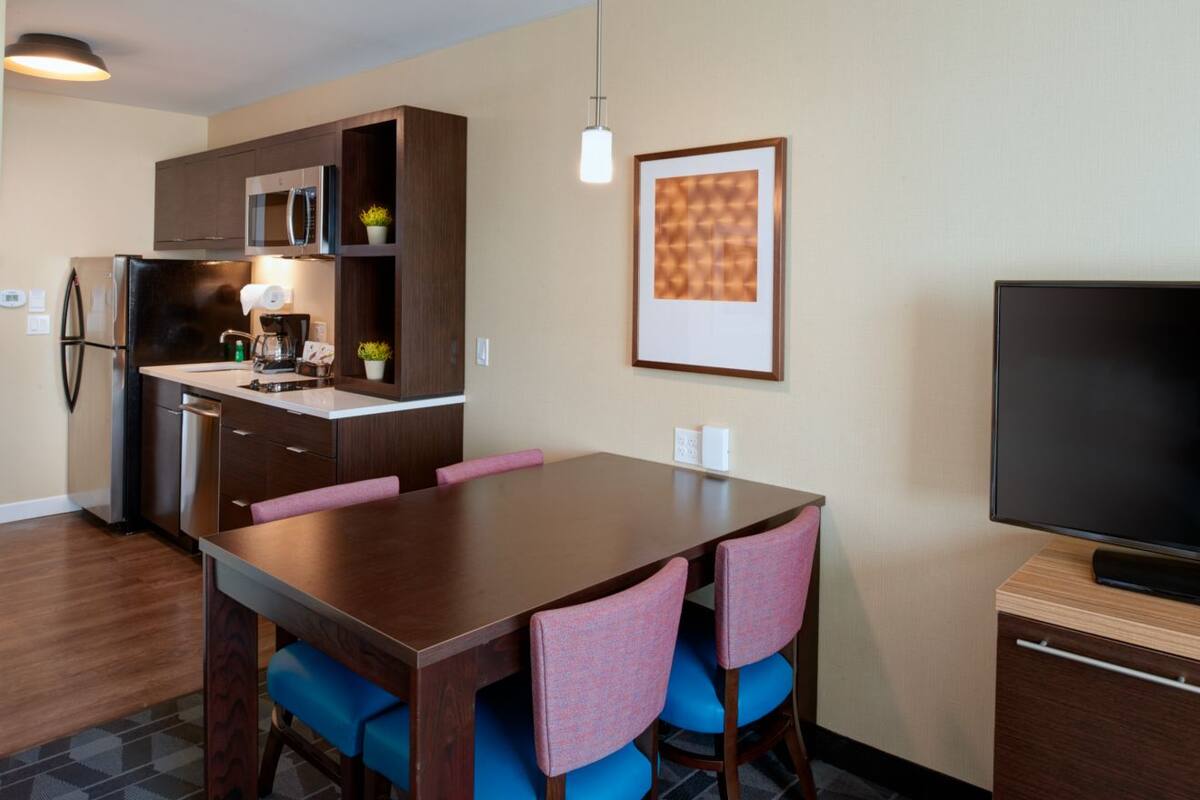 A modern kitchen with dark wooden cabinets, a stainless steel refrigerator, and a built-in microwave. There is a dining table with blue upholstered chairs and a flat-screen TV on a wooden stand.