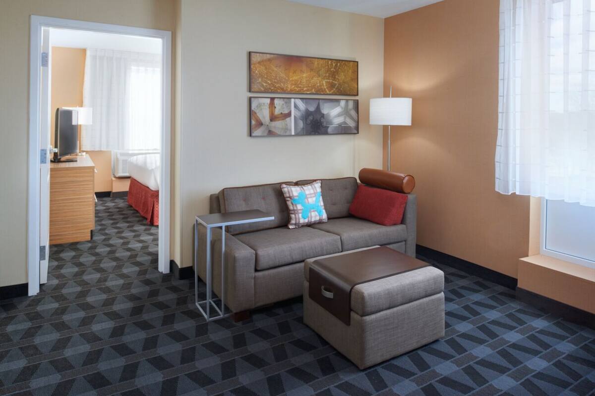 A hotel room with a sofa, ottoman, and a TV on a wooden dresser.