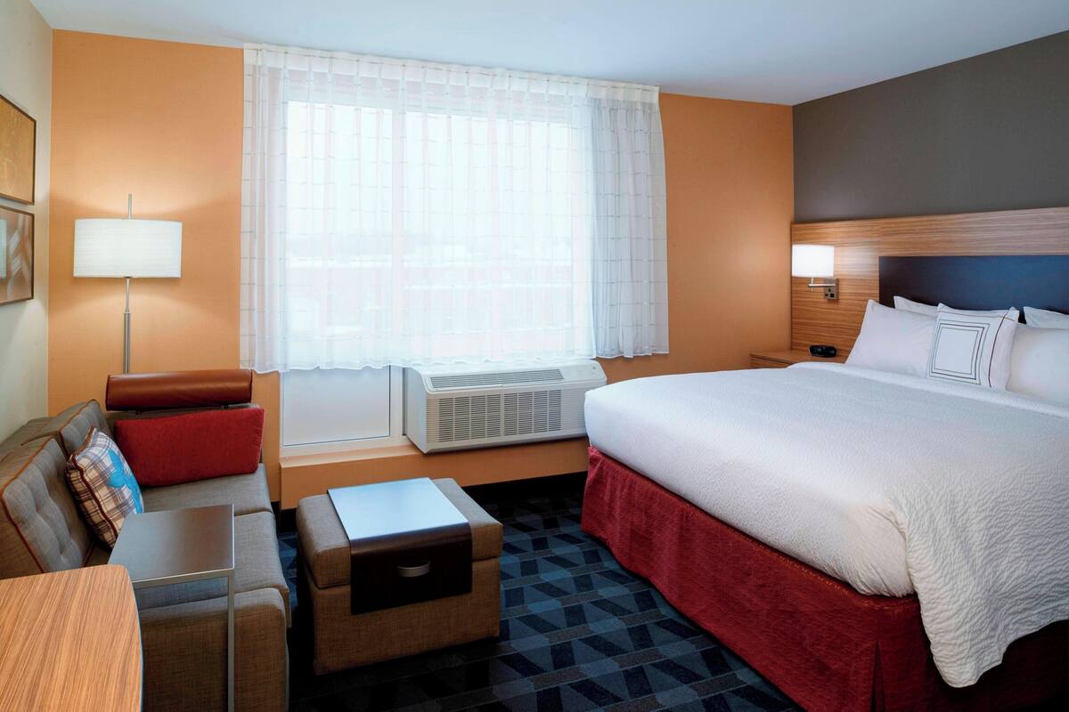 A hotel room with a bed, a desk, a chair, a sofa, a lamp, and a window with curtains.