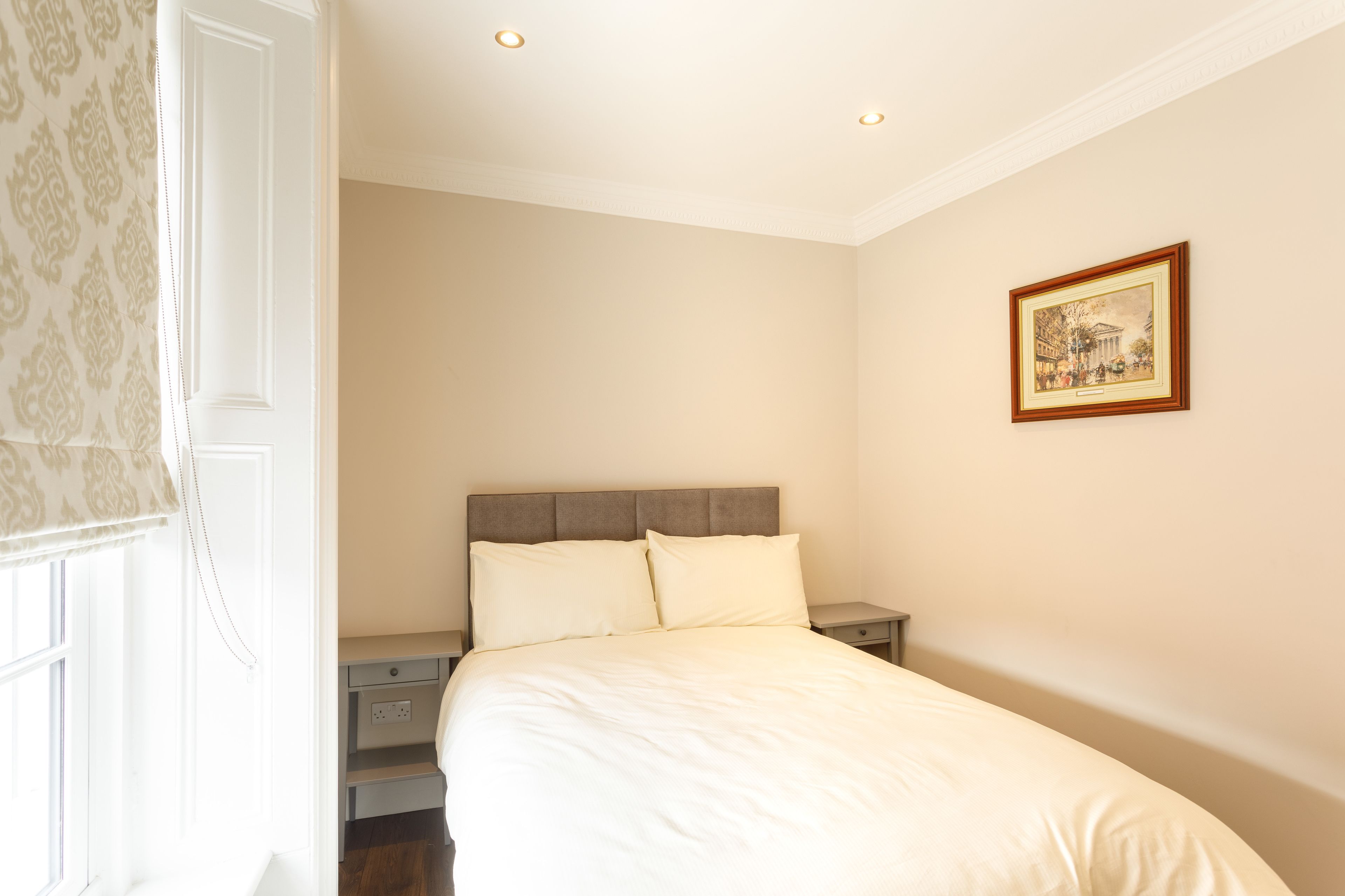 A bedroom with a large bed, a nightstand, a small drawer, and a framed picture on the wall.