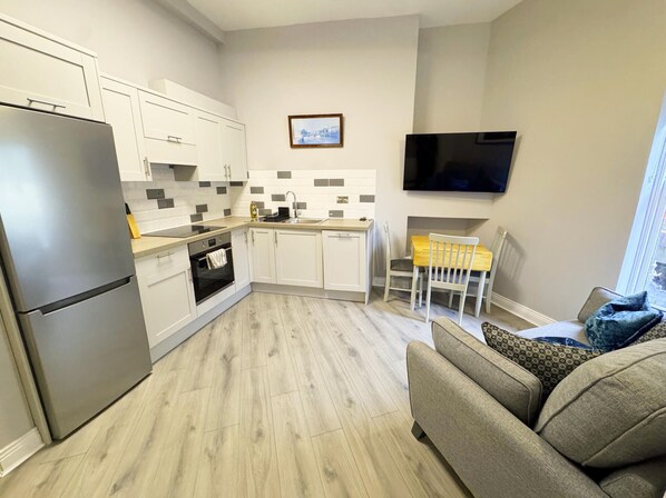 A modern kitchen with white cabinets, a built-in oven, and a grey refrigerator. There is a dining area with a wooden table and chairs, and a flat-screen TV mounted on the wall.