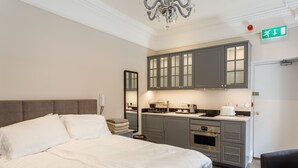 Un dormitorio con una cama grande, una mesita de noche, un espejo y una kitchenette con armarios y electrodomésticos.