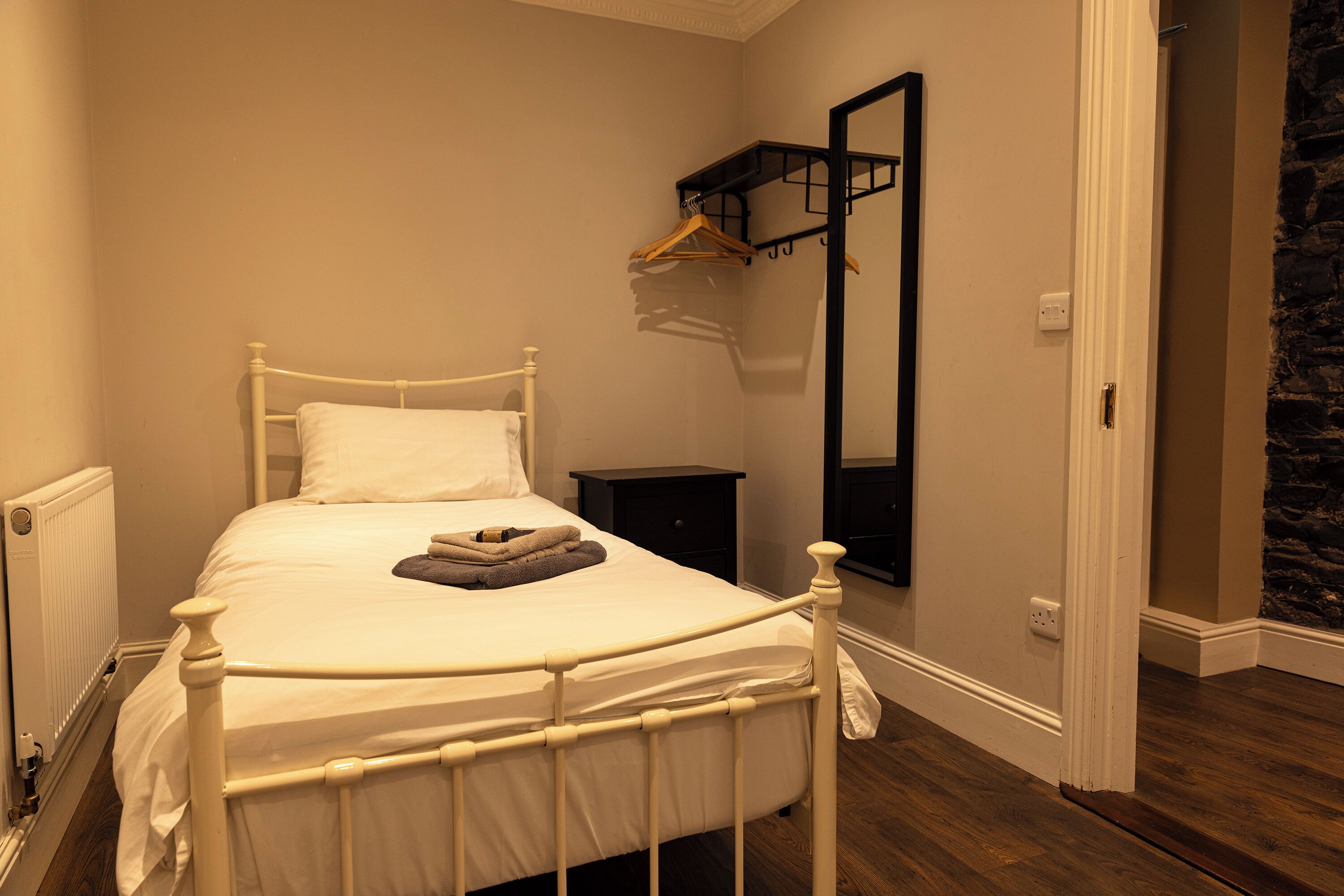 A neatly made bed with a white headboard, a bedside table, a wall-mounted mirror, and a wooden floor.