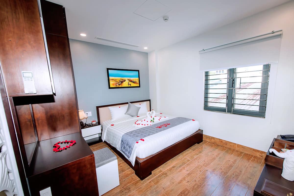 A hotel room with a wooden bed, a nightstand, a desk, and a window with blinds.