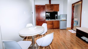 A compact kitchen with wooden cabinets, a refrigerator, and a dining area with a glass table and white chairs. - My Family (Da Nang)
