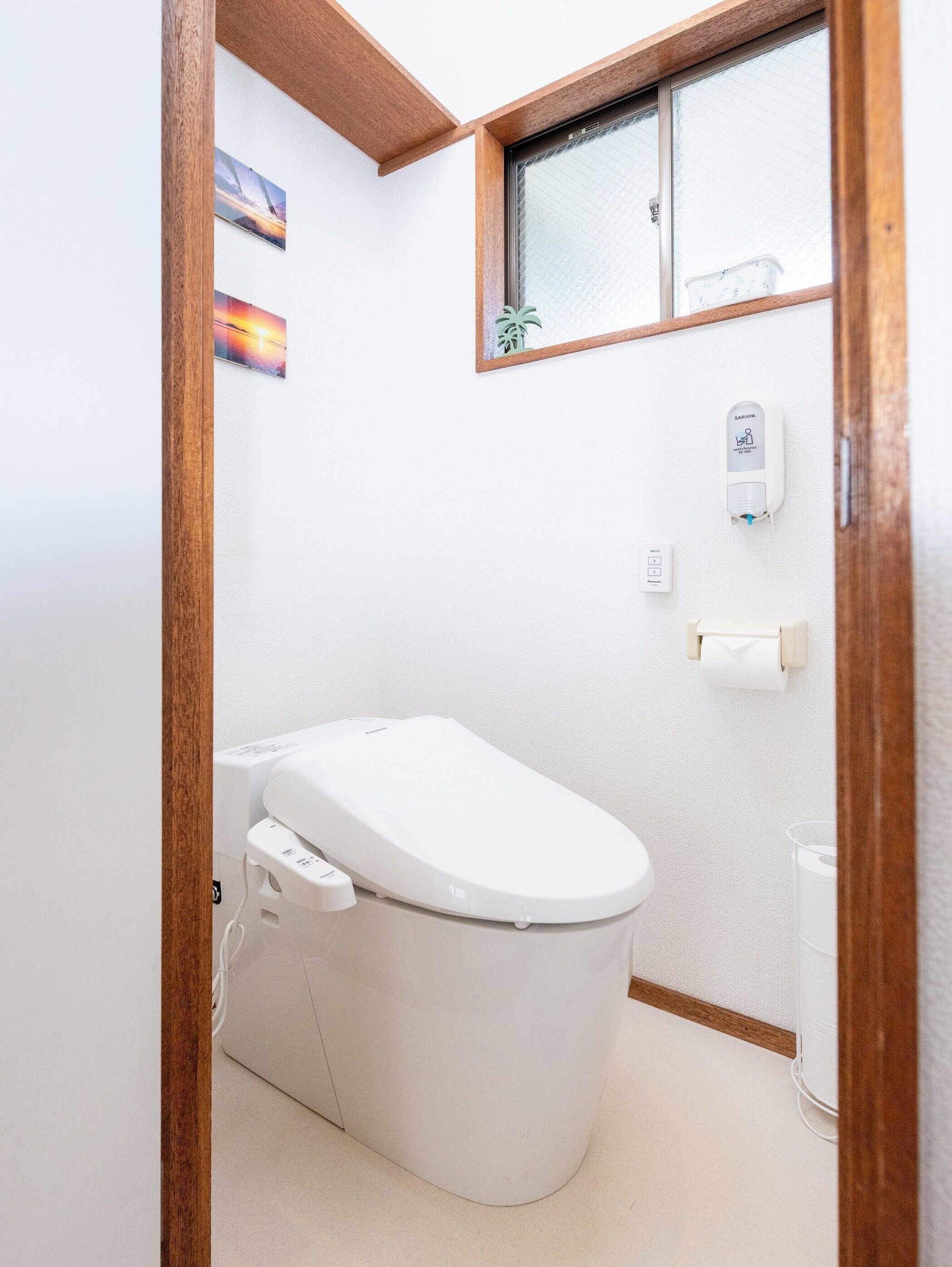 A modern bathroom with a toilet, a hand sanitizer dispenser, and a window with a view of a palm tree.