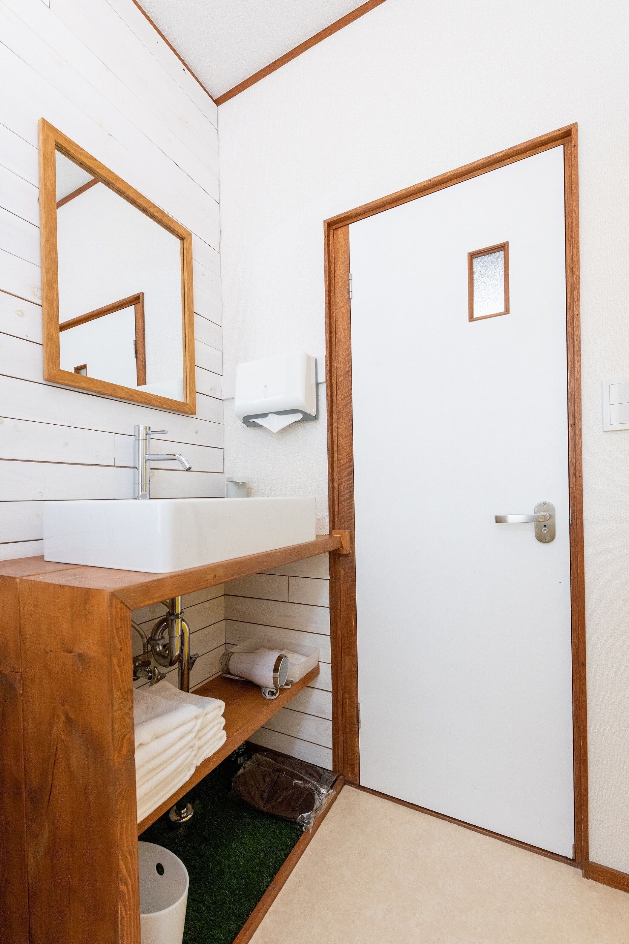 Une salle de bain avec un meuble-lavabo en bois, un lavabo blanc, un miroir et une porte dotée d’une petite fenêtre.