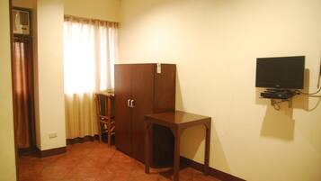 A room with a wardrobe, a small table, a television mounted on the wall, and a window with curtains.