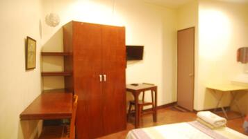 Un dormitorio con cama, un armario de madera, un escritorio, una silla, una mesita y un televisor en la pared.