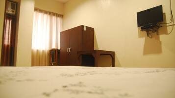 A hotel room with a bed, a wooden wardrobe, a small table, a television mounted on the wall, and a window with curtains.