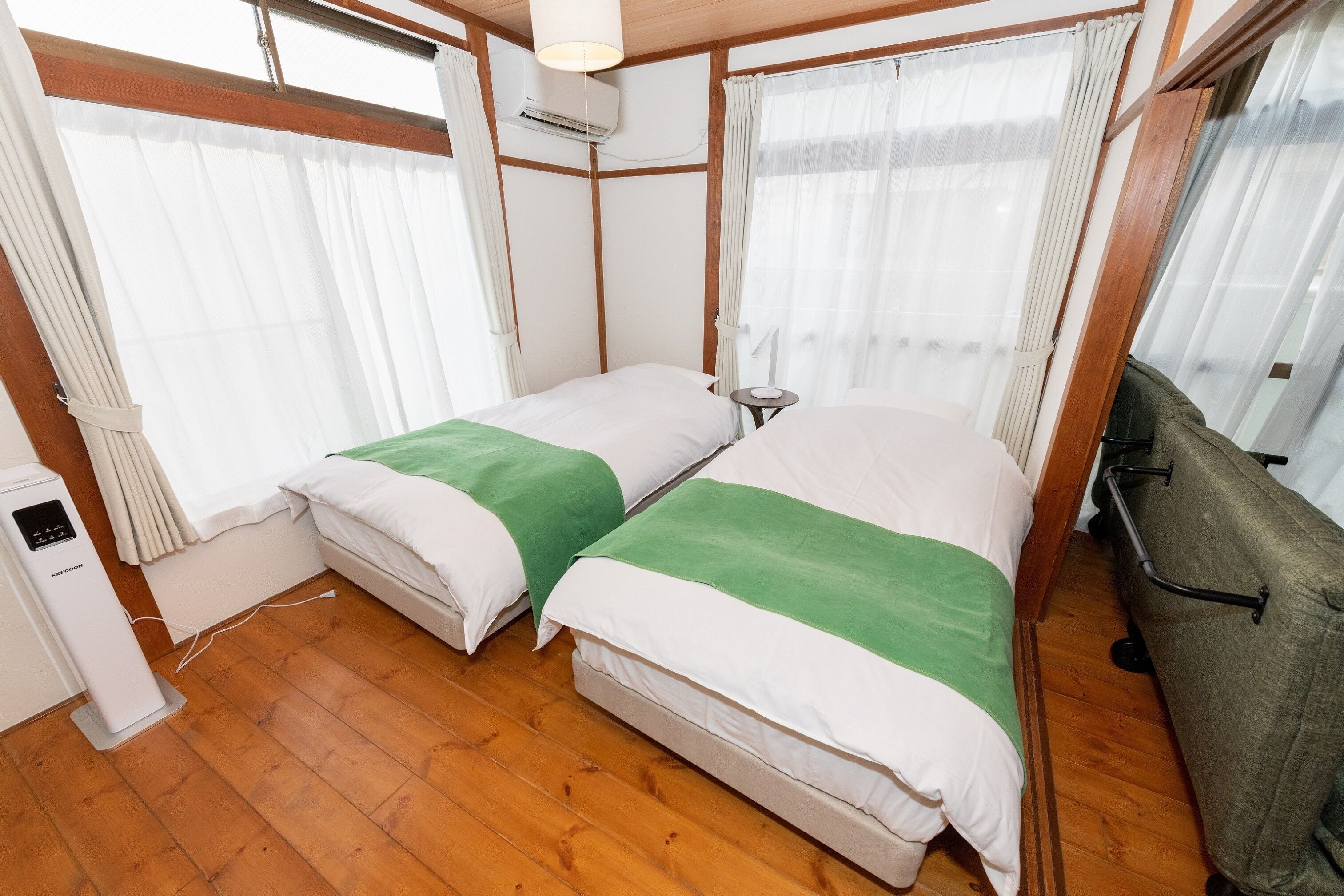 A compact hotel room with two beds, wooden flooring, a small bedside table, a heater, and a window with curtains.