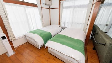 A compact hotel room with two beds, wooden flooring, a small bedside table, a heater, and a window with curtains.