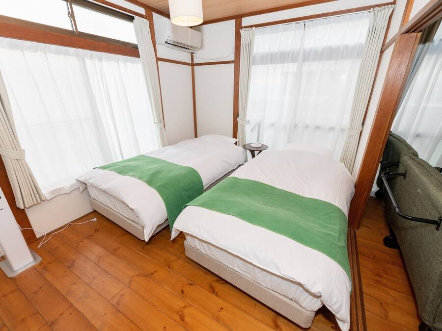 A compact hotel room with two beds, wooden flooring, a small bedside table, a heater, and a window with curtains.