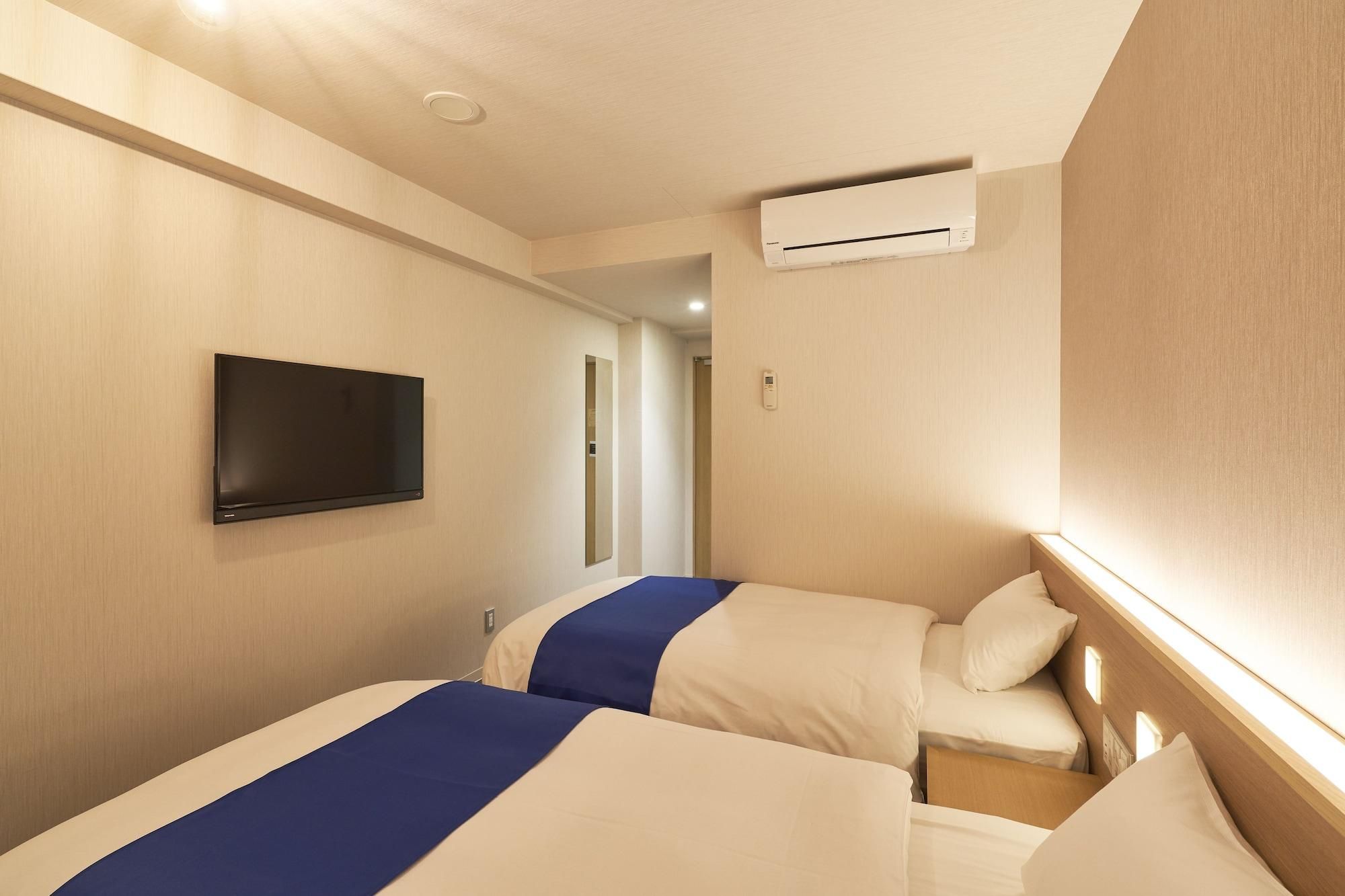 A hotel room with two beds, a flat-screen TV mounted on the wall, an air conditioning unit, and a bedside table with a lamp.