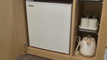 A compact refrigerator in a wooden cabinet with a glass shelf above containing two white mugs and a white appliance below.