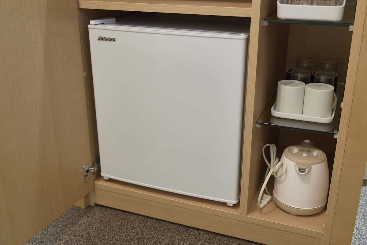 A compact white refrigerator in a wooden cabinet with shelves above containing cups and a kettle.