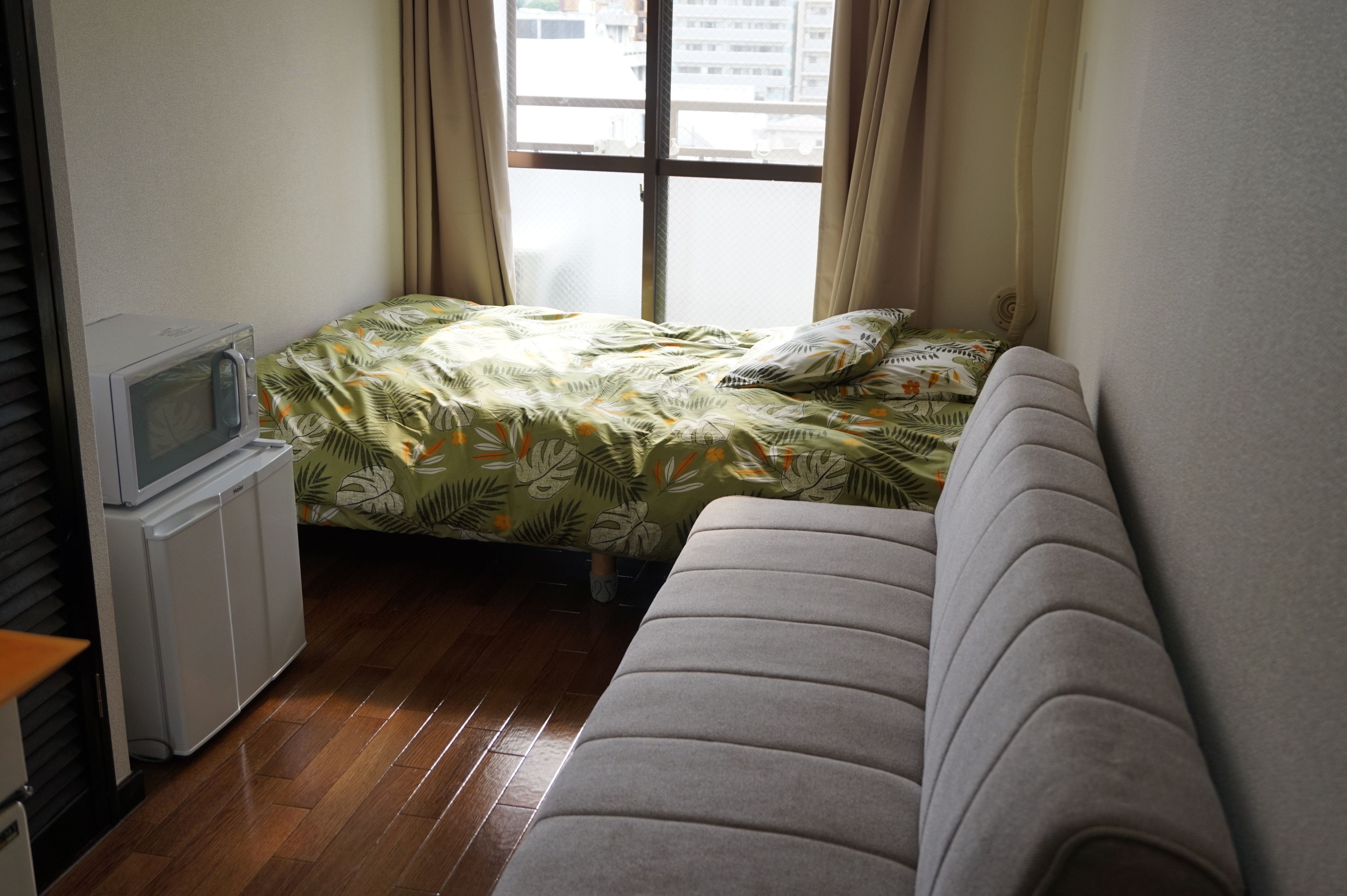 Um quarto pequeno e bem iluminado, com uma cama, um sofá e uma televisão sobre um suporte.