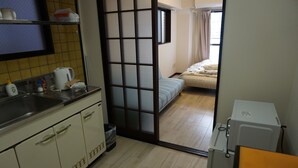 Eine kleine Küche mit Spüle, Wasserkocher und Schränken. Eine Schiebetür führt in ein Schlafzimmer mit Bett und Fenster.