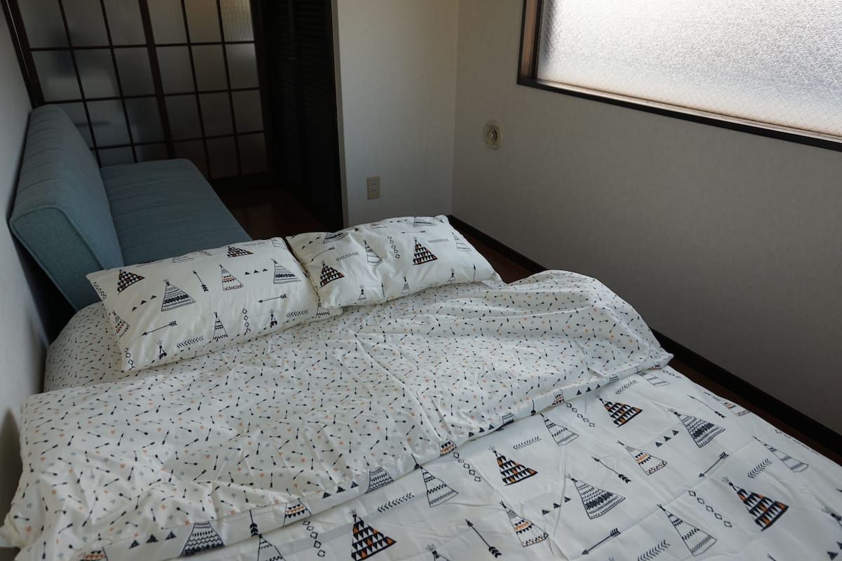 Ein Bett mit weißer Bettwäsche mit geometrischem Muster, einem blauen Kissen und einem Fenster mit Jalousien.