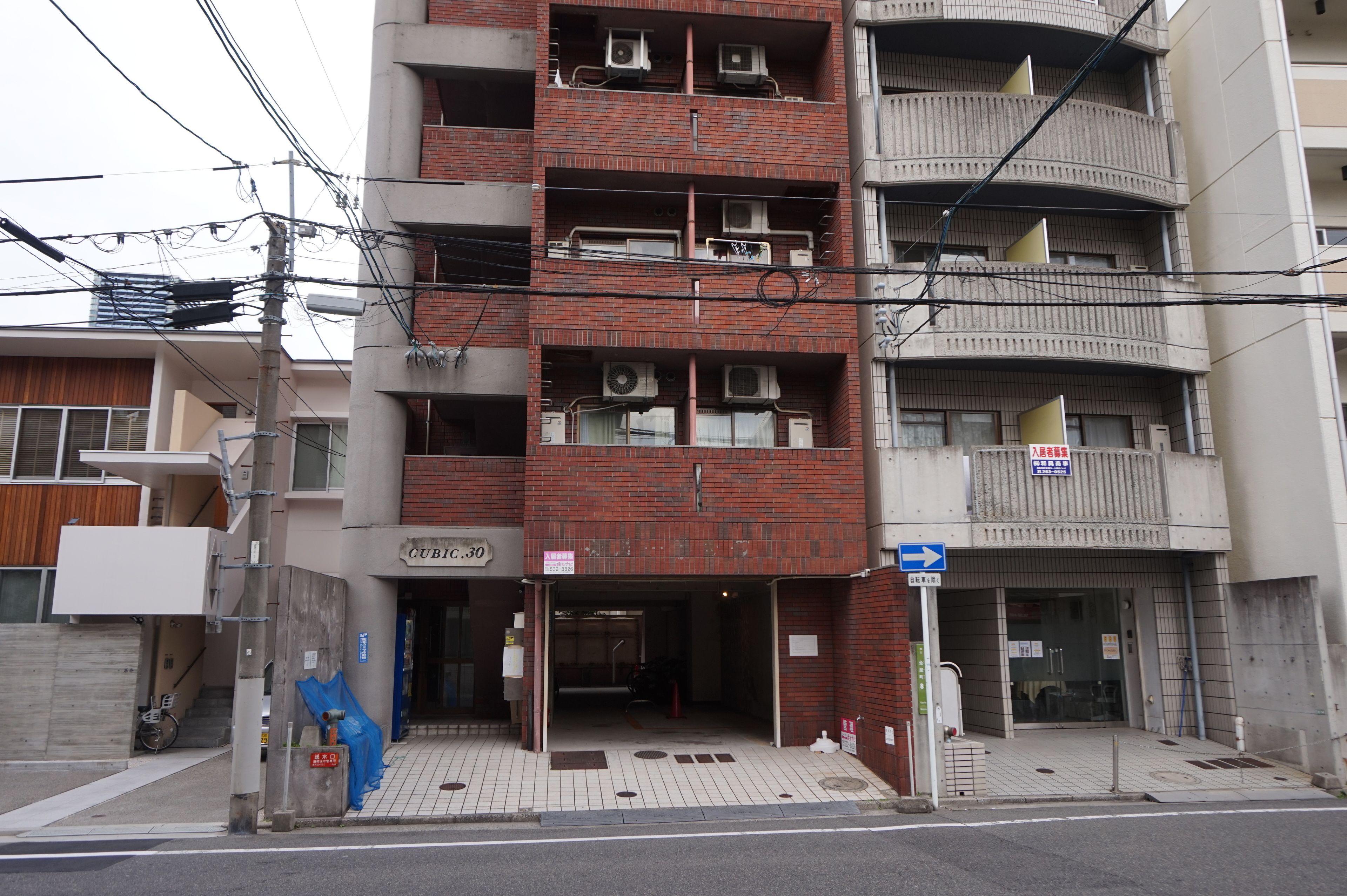 Um edifício com vários andares, entrada no nível da rua, aparelhos de ar condicionado e uma placa indicando uma área comercial ou de serviços.