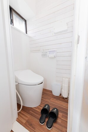 A small, clean bathroom with a white toilet, a wooden floor, and a pair of black slippers. - Blue Lagoon (Kamakura)