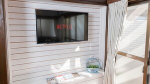 A room with a mounted television displaying the Netflix logo, a small table with magazines, and a window with curtains. - Blue Lagoon (Kamakura)