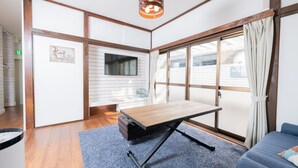 A room with a foldable table, a sofa, a TV mounted on the wall, and a large window with curtains. - Blue Lagoon (Kamakura)