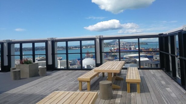 A rooftop terrace with wooden tables and benches, large windows offering a view of the sea and port. - Brand New Apartment on Anzac Ave (Auckland)