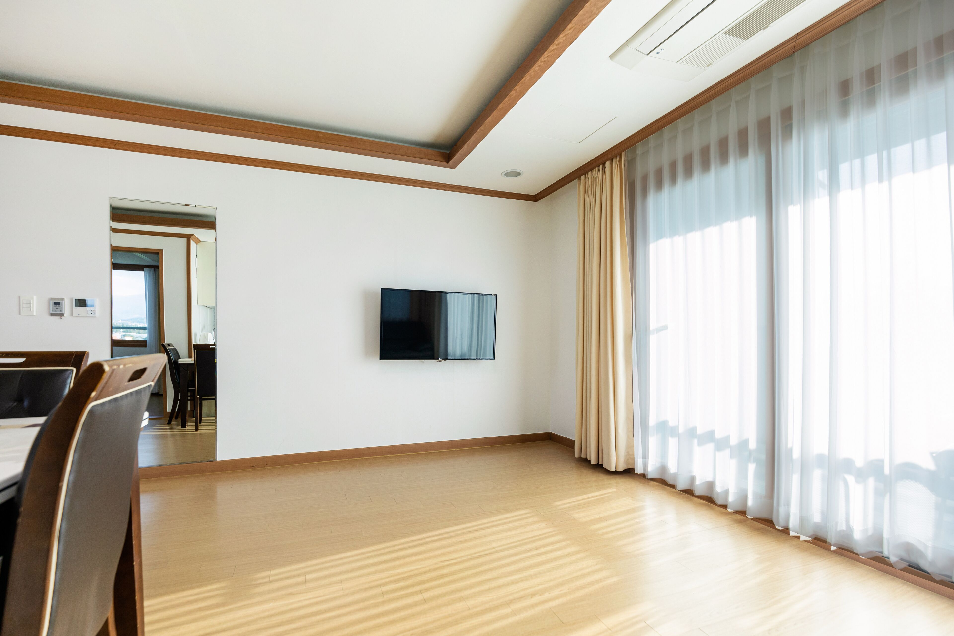 A spacious room with a wooden floor, a flat-screen TV mounted on the wall, and large windows with sheer curtains.