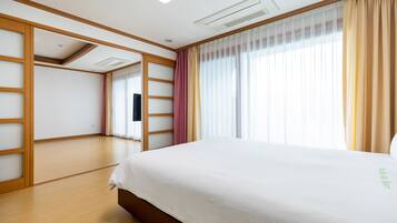 Una habitación de hotel con una cama grande, suelo de madera, ventilador de techo y un amplio ventanal con cortinas.