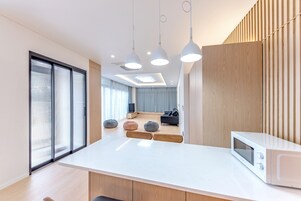 Una cocina moderna con encimera blanca, microondas, luces colgantes y puerta corredera de vidrio hacia una sala de estar.