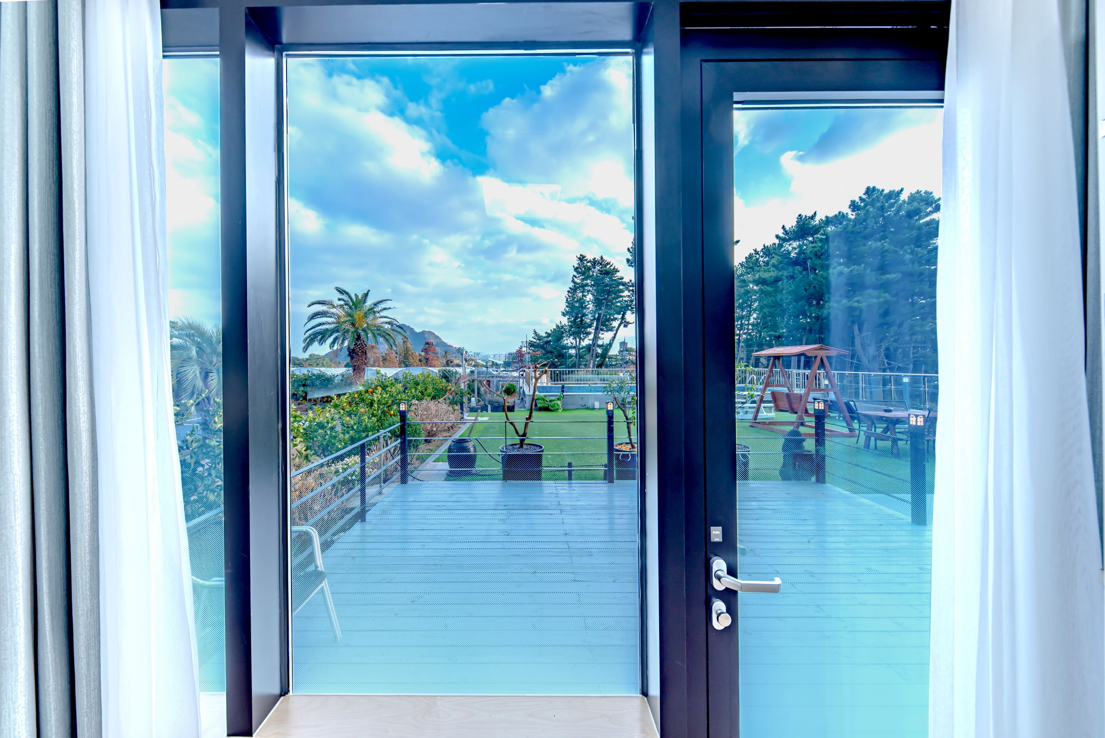 A view through a glass door to an outdoor deck with a metal railing, a palm tree, and a clear sky.
