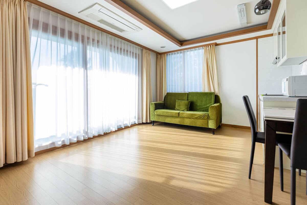 Una habitación amplia con suelo de madera, un sofá verde, una mesa de comedor con sillas y ventanas grandes con cortinas traslúcidas.