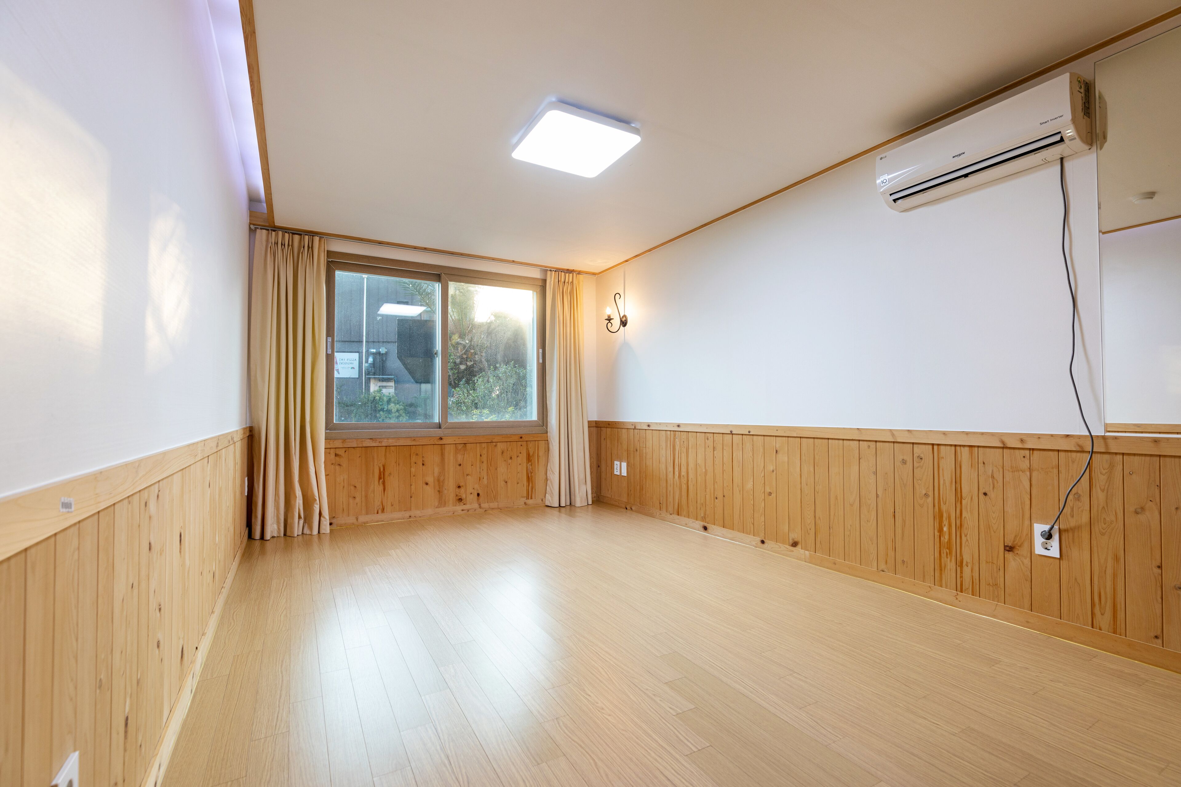 Een kamer met houten lambrisering, een groot raam met gordijnen, een airconditioning en een plafondlamp.