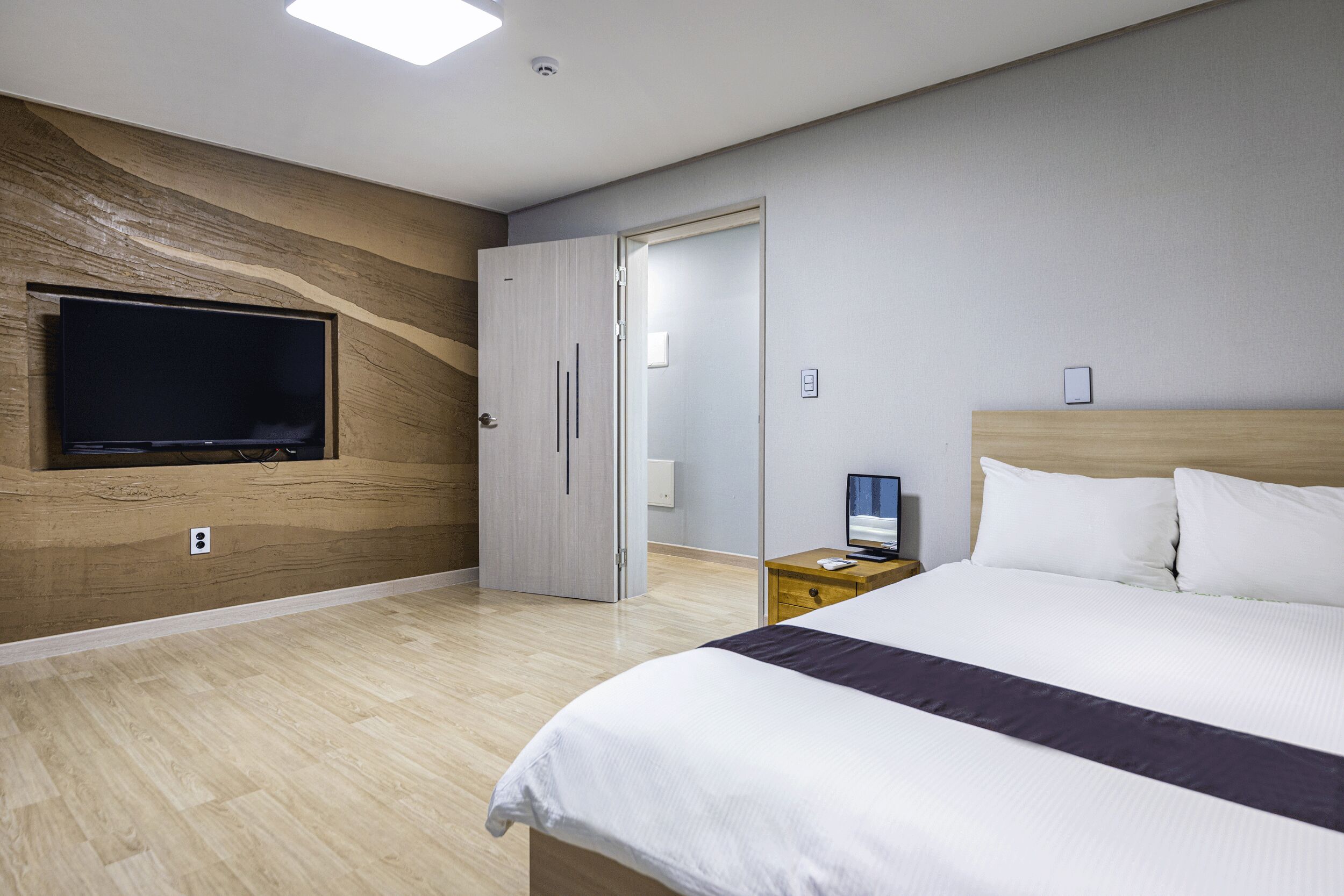 A modern hotel room with a large bed, a flat-screen TV mounted on the wall, and a wooden accent wall.