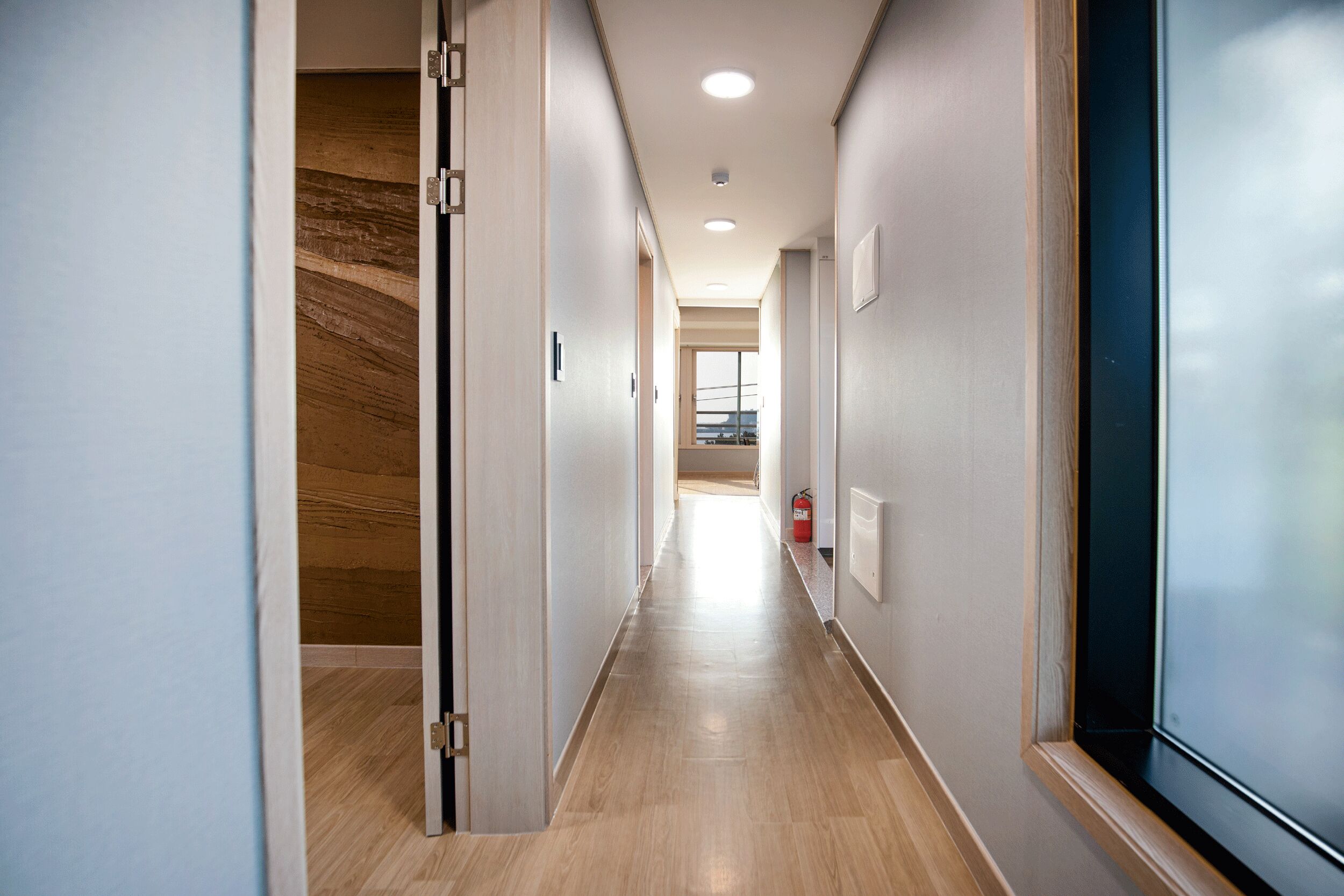 A modern hallway with wooden flooring, recessed ceiling lights, and a fire extinguisher on the wall.