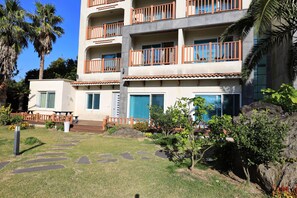 A multi-story hotel with balconies, a garden area with stepping stones, and palm trees. - The Bay Jeju Resort (Seogwipo)