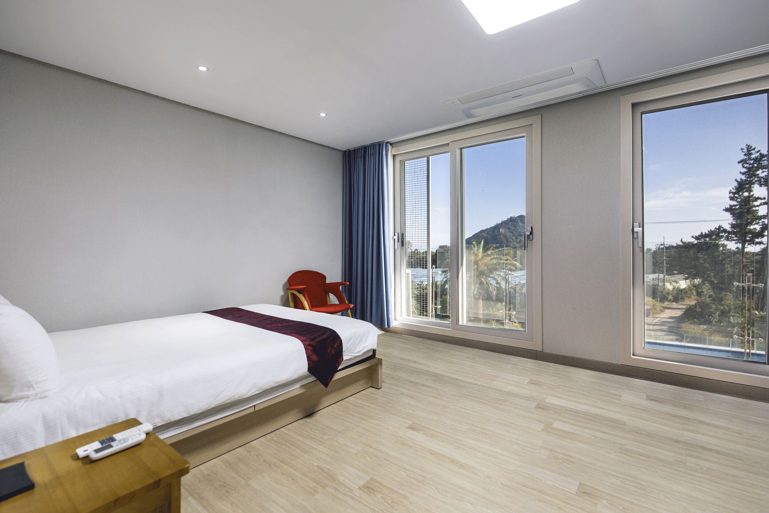 A modern hotel room with a bed, a chair, a nightstand, and large windows offering a view of the outdoors.