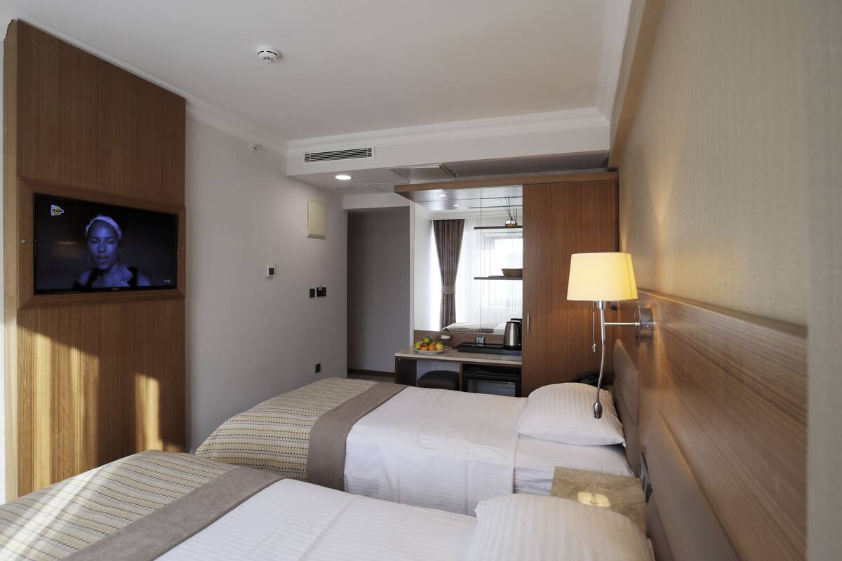 A hotel room with two beds, a TV mounted on the wall, a desk with a mirror, and a lamp.