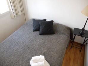 A neatly made bed with a grey patterned bedspread and two black pillows. A white towel is placed at the foot of the bed. There is a small black table with a lamp beside the bed. - Santa Fe & Carranza By Foreign In Baires (Buenos Aires)