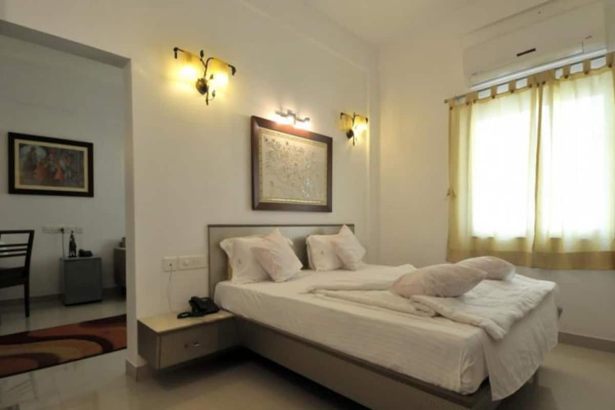 A hotel room with a large bed, bedside tables, a desk, and a chair. There are wall-mounted lights and a window with curtains.