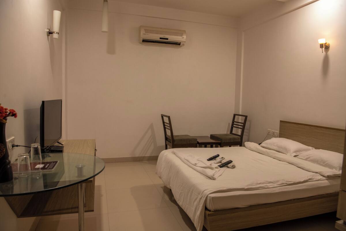A hotel room with a bed, a glass table, a chair, a TV, and an air conditioning unit.
