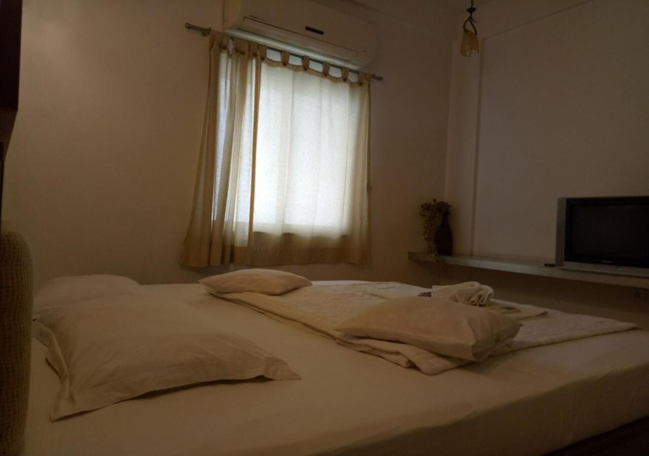 Uma cama de solteiro com almofadas e cobertor, uma janela com cortinas, uma plantinha e uma televisão sobre um suporte.