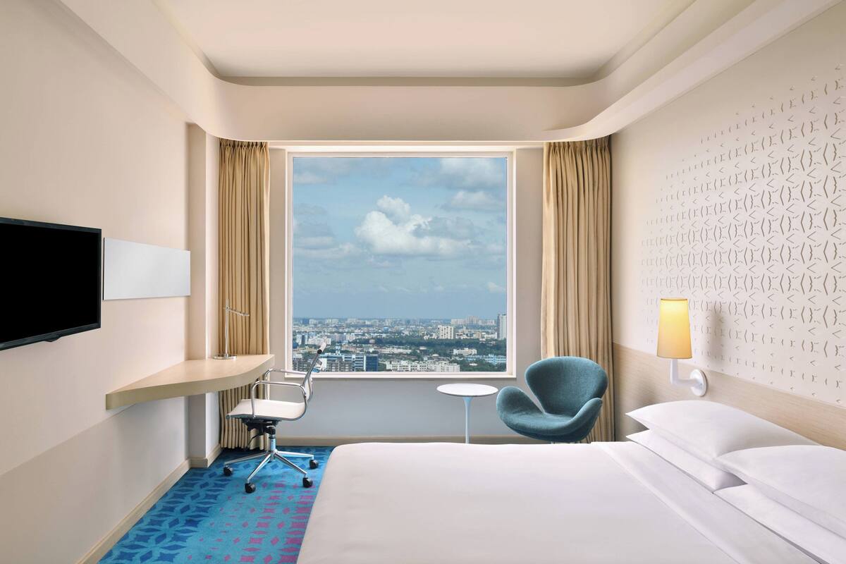 Ein Hotelzimmer mit Bett, Schreibtisch mit Stuhl, Fernseher, einem kleinen Tisch, einer Lampe und Blick auf die Stadt durch ein großes Fenster.