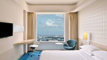 Habitación de hotel con cama, escritorio con silla, televisor, mesita pequeña, lámpara y vista de la ciudad a través de un ventanal amplio.