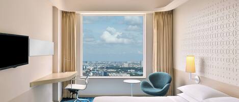 A hotel room with a bed, a desk with a chair, a TV, a small table, a lamp, and a view of the city through a large window.