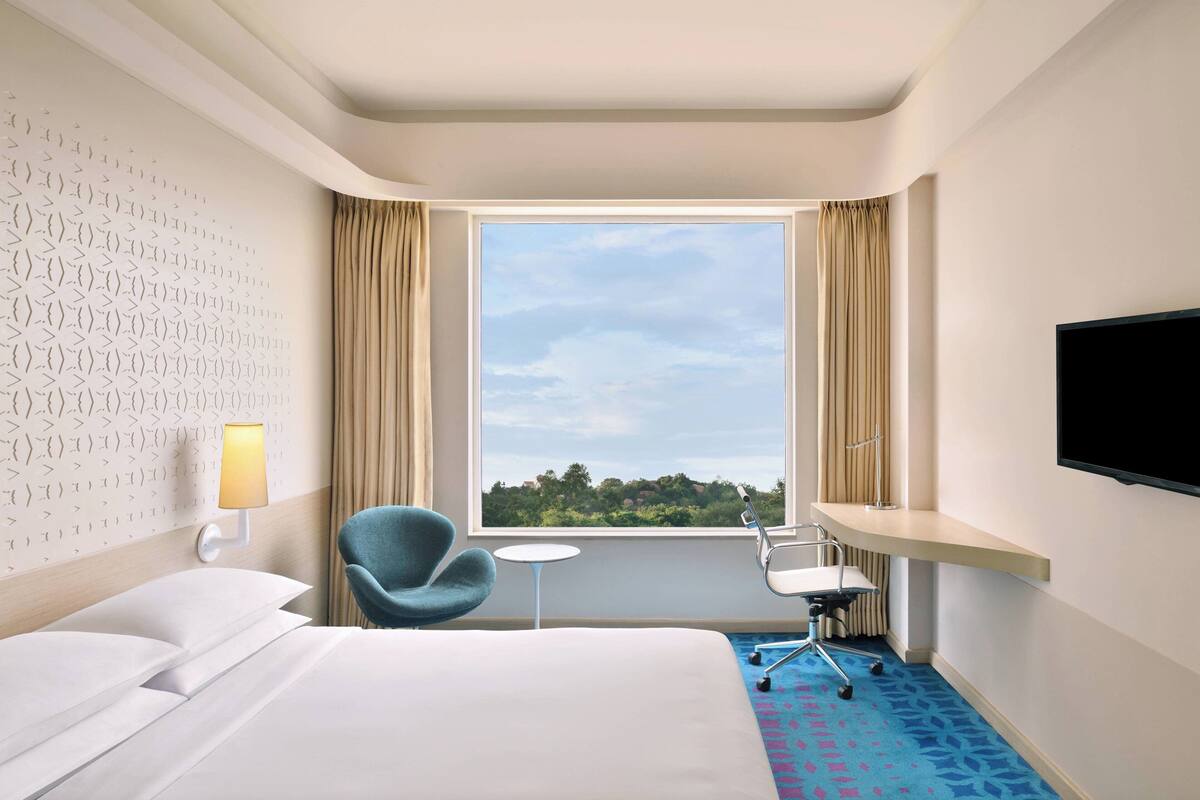 Ein Hotelzimmer mit Bett, Sessel, kleinem Tisch, Fernseher und Fenster mit Blick ins Grüne.